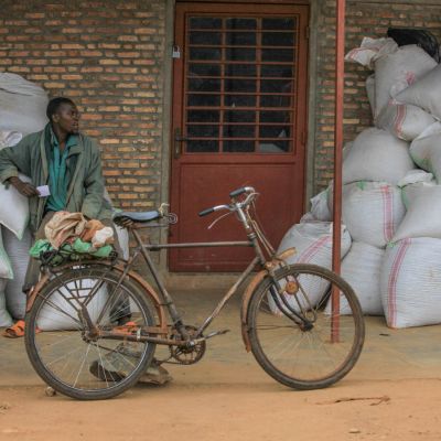 Bags of parchment coffee are piled up on either side of the door outside the office at the Murambi green coffee washing station in Kiganda, Burundi, and an old bicycle is parked against a pole. 