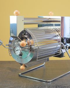 Angled frontview of the Quest M6 electric coffee roaster. The barrel faces forward with a chaff collector below and an air intake tube above. 