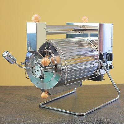 Angled frontview of the Quest M6 electric coffee roaster. The barrel faces forward with a chaff collector below and an air intake tube above. 